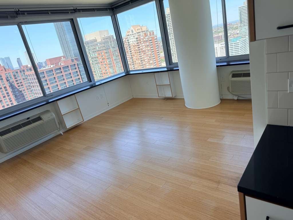 Empty high-rise apartment interior with hardwood floor, large windows overlooking urban cityscape, white column, and minimalist furnishings