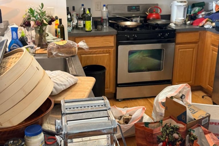 Cluttered kitchen with wooden cabinets, stainless steel oven, pasta maker, and various items scattered on countertops and floor