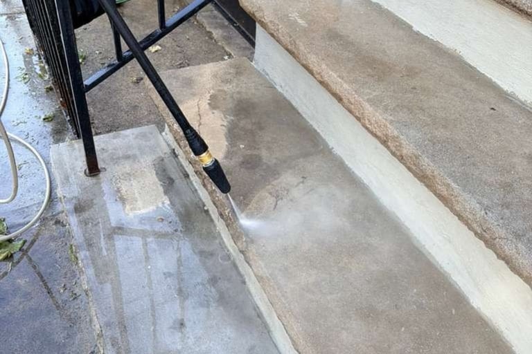 Pressure washer spraying water on concrete surface with chair and hose visible in background
