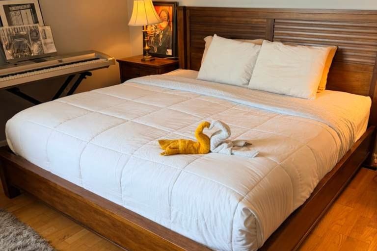 A wooden bed with a white comforter and gray pillows, featuring a yellow stuffed animal on the mattress, in a warmly lit bedroom.