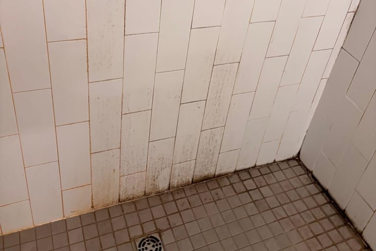 Corner of a tiled bathroom shower with white subway tiles on walls and small brown floor tiles, showing water stains and discoloration
